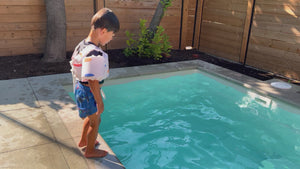 Vidéo d'un garçon vue de dos saute dans une piscine creusée moderne avec son flotteur blanc avec motifs de surfs orangé, corail et bleu. Après son saut, il remonte à la surface et nage au rebord de la piscine.